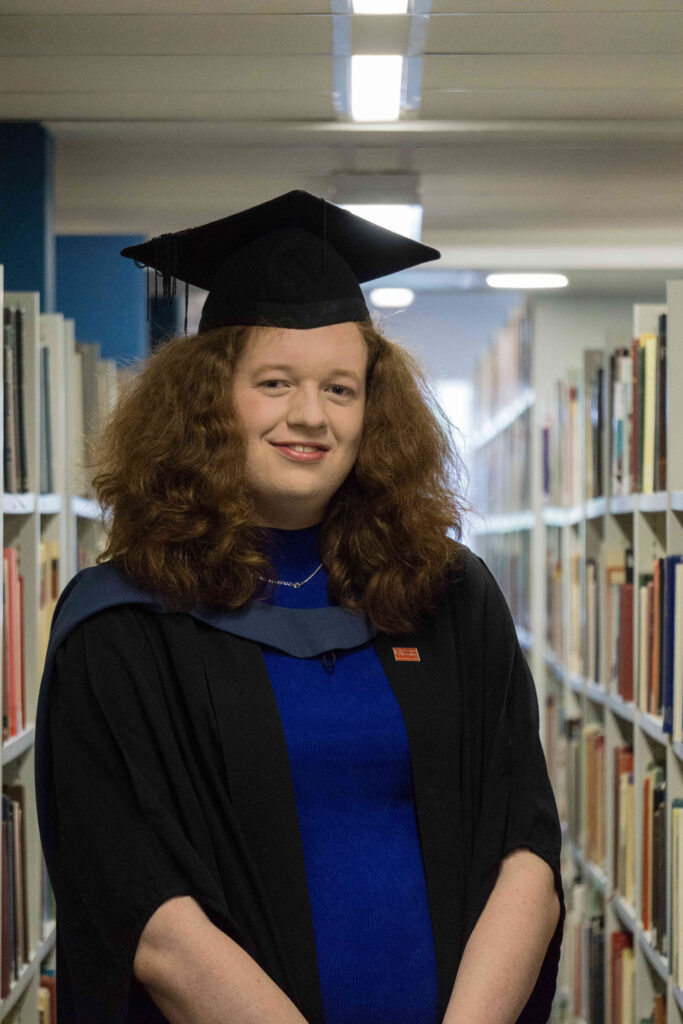 Olivia, stood inbetween libary bookshelfs. Olivia is wearing a blue dress with graduation gown, hood and morterboard